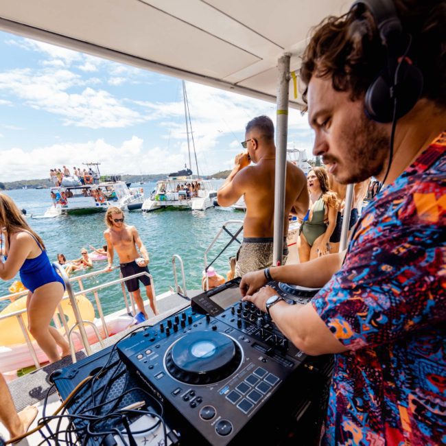 A DJ wearing headphones performs on a luxury yacht, playing music using a mixing console while people on an adjacent boat enjoy the sunny day and water activities.