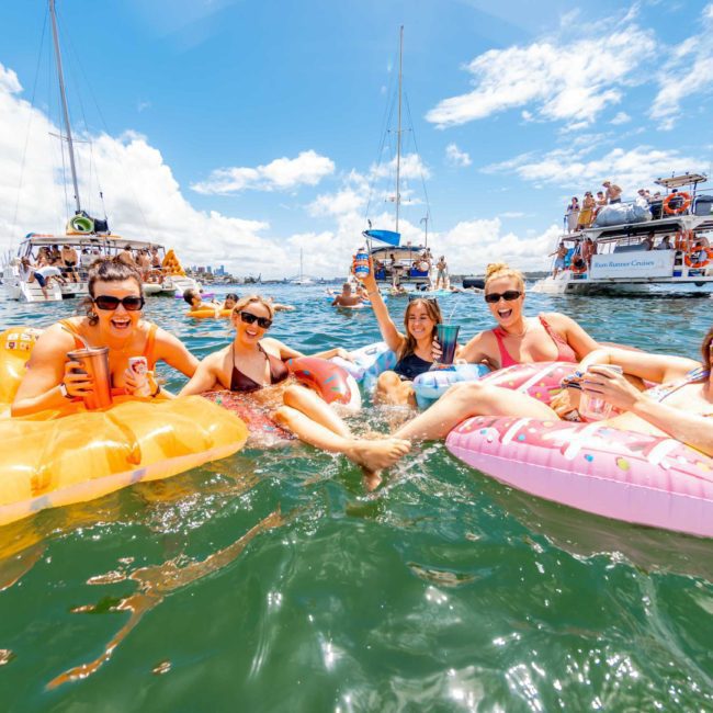Five people float on inflatable loungers in a body of water, holding drinks. Boats with more people are visible in the background under a bright, sunny sky. It's the perfect setting for a Catamaran party Sydney is known for.