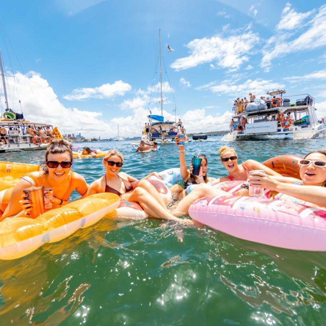 A group of people relax on inflatable floats in the water with several luxury yachts in the background under a blue sky with scattered clouds, enjoying a private yacht charter on Sydney Harbour.