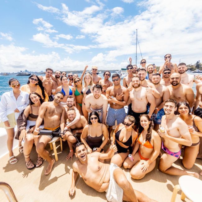 A large group of people in swimwear pose together on a boat under a sunny sky. The background features water and a distant shoreline, the perfect setting for a Sydney boat party hire.