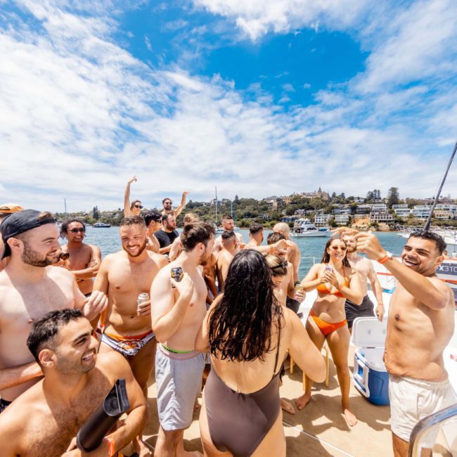A group of people in swimwear gather on a boat under a partly cloudy sky, enjoying what appears to be a social event or private yacht charter Sydney Harbour.