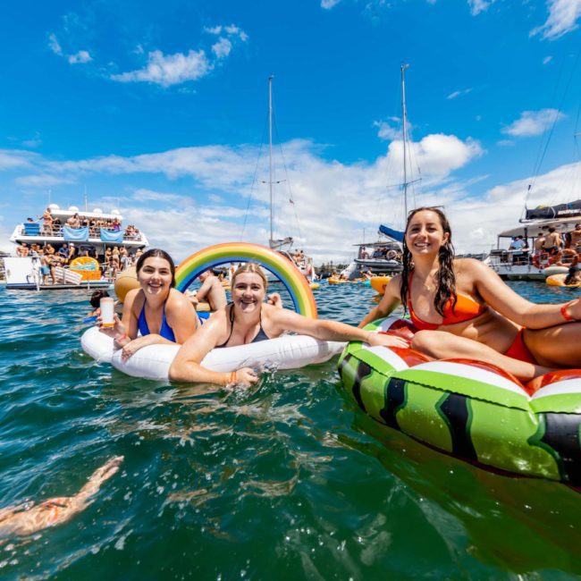 Three people in swimsuits are floating on inflatables in a lake with boats and other people in the background on a sunny day, enjoying what looks like the perfect catamaran party Sydney has to offer.