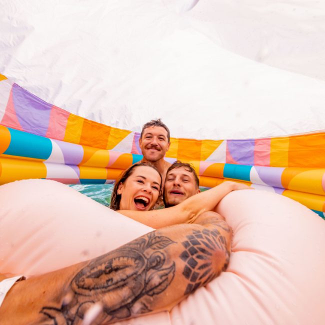 Four people smiling inside a colorful inflatable structure with water during a festive Sydney boat party hire. A tattooed arm is visible in the foreground, adding to the vibrant atmosphere.