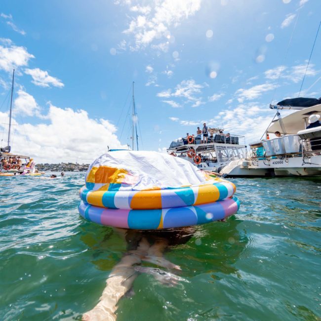 A person floats on a multicolored inflatable ring in the water near several anchored boats under a bright, sunny sky, enjoying the vibrant atmosphere of a private yacht charter in Sydney Harbour.