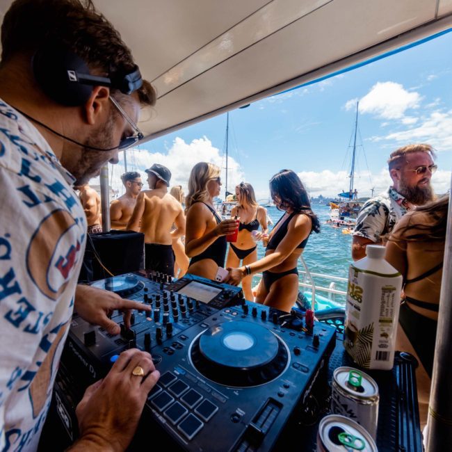 DJ playing music on a boat surrounded by people in swimwear. The background shows a sunny day with other boats on the water, making it an unforgettable catamaran party Sydney experience.