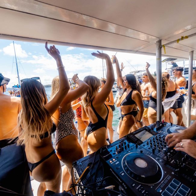 People in swimwear dancing on a luxury yacht hire Sydney with a DJ playing music. The scene includes clear skies and water in the background.