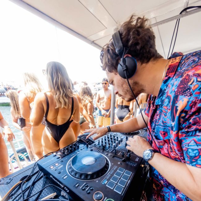 A DJ wearing headphones and a colorful shirt operates a mixing console at a beach party with people in swimwear dancing in the background, reminiscent of the energy found in Sydney boat party hire events.