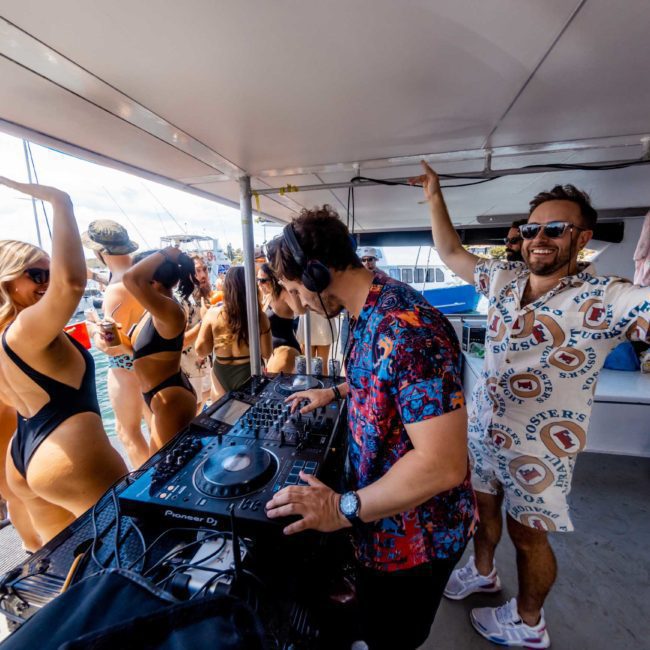 A DJ is playing music on a boat while people in swimwear are dancing and enjoying a sunny day at a Catamaran party Sydney.