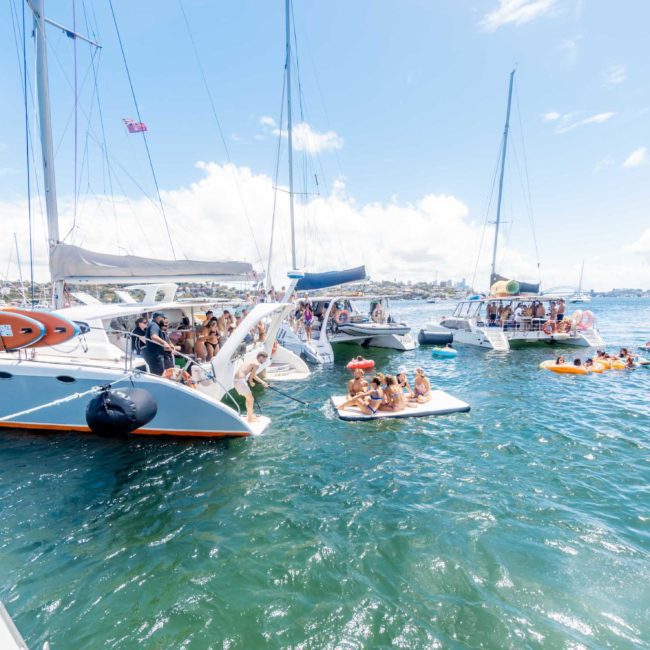 Several sailboats and small crafts are anchored closely together in a body of water on a sunny day, with people swimming and socializing on the boats and inflatable rafts. Experience the ultimate catamaran party Sydney has to offer, perfect for those looking to enjoy a lively day on the water.