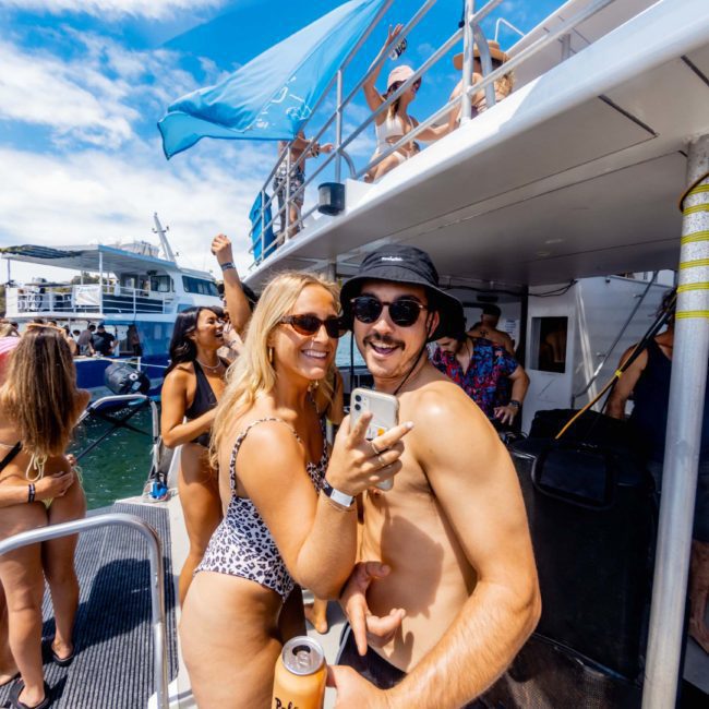 People on a boat enjoying a sunny day. A woman in a leopard print swimsuit and a man in sunglasses and a hat pose for a photo. Other people in swimwear are around them, having fun on and around the boat, taking full advantage of their luxury yacht hire Sydney experience.