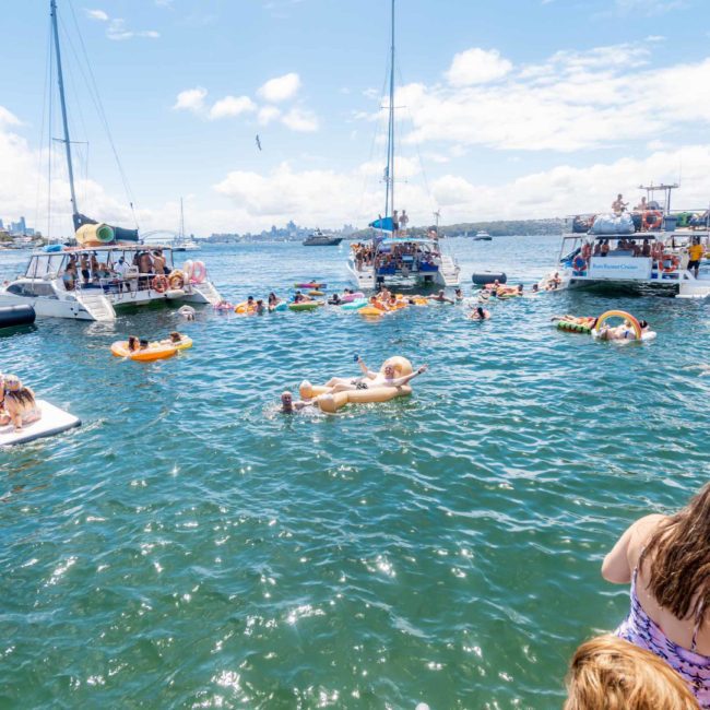 People swimming and relaxing on floaties at a Catamaran party in Sydney, enjoying a boat party gathering on a sunny day, surrounded by several boats in the water.