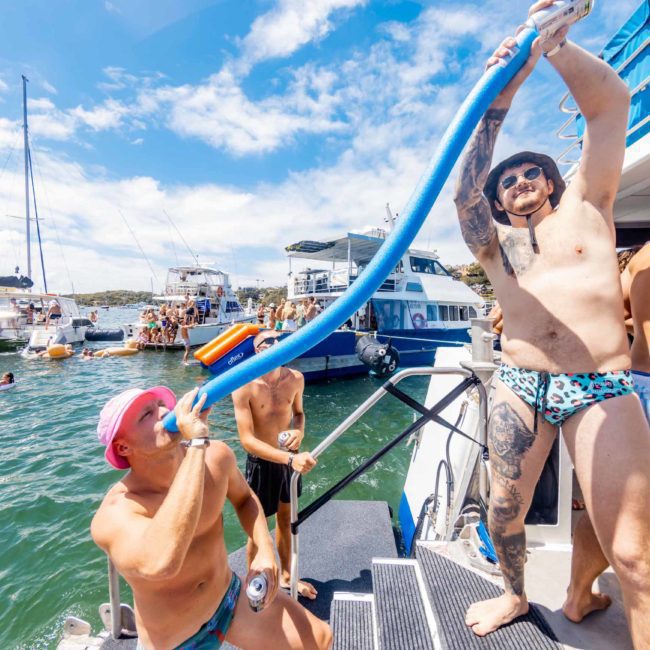 At a lively nautical gathering, two men are on a boat, one drinking from a tube held by the other. In the background, other boats and people swimming enhance the festive atmosphere. Experience such moments with a private yacht charter on Sydney Harbour for an unforgettable adventure.