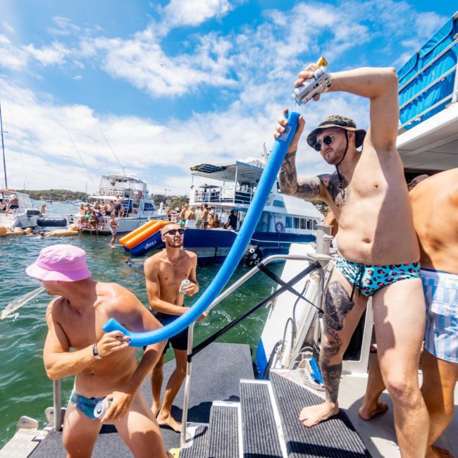 People on a boat enjoying a sunny day; one person is on the stairs holding a pool noodle, another wearing a hat is pulling it, and others are in swimwear. In the backdrop, boats drift on Sydney Harbour, perfect for luxury yacht hire or private yacht charters.