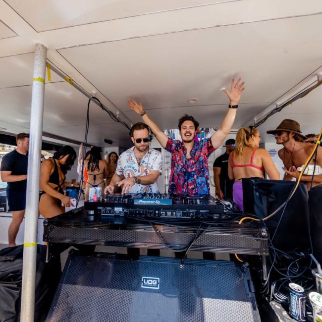Two DJs perform on a luxury yacht with a crowd of people dancing and socializing around them.