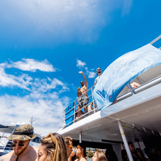 People enjoying a sunny day on a multi-level boat in Sydney Harbour, with some standing on the upper deck waving. The boat's banner reads "SOCIAL." Bright blue sky and a few clouds in the background. Perfect setting for luxury yacht hire Sydney or private yacht charter events.