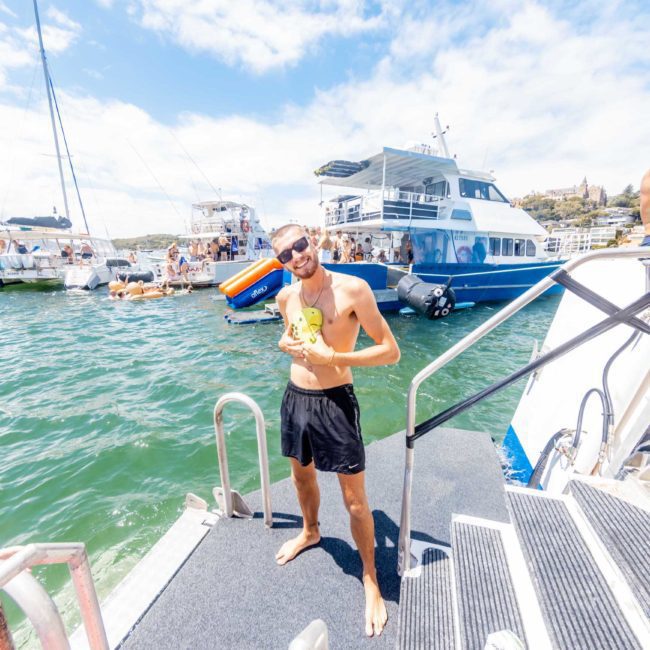 A man in black shorts stands on the step of a boat holding a toy while smiling, with other boats and people visible in the background on a sunny day. Perfect setting for a Sydney boat party hire or private yacht charter Sydney Harbour event.
