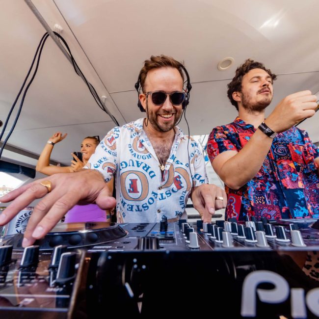 A man in a colorful shirt and sunglasses is DJing at a mixer on a luxury yacht hire in Sydney, while another man in a patterned shirt stands beside him. People are in the background under a canopy.