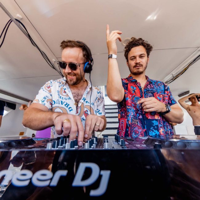 Two DJs perform on a private yacht charter in Sydney Harbour, with one wearing headphones and adjusting the controls, while the other gestures with his hand. Onlookers in casual attire are seen in the background.