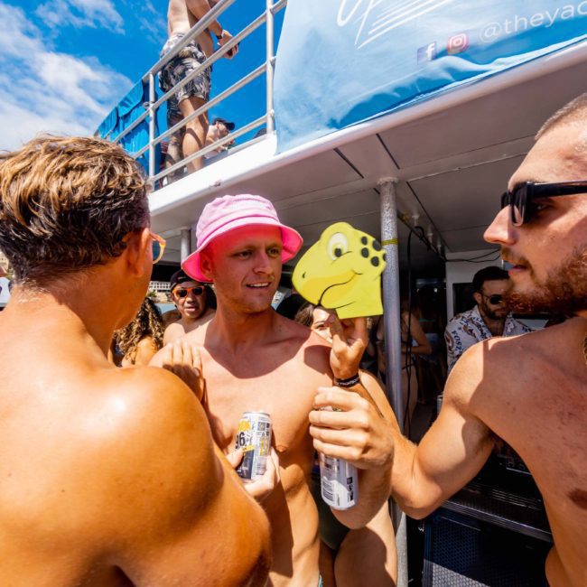 A group of shirtless men enjoy a lively Catamaran party in Sydney. One holds a cutout of a cartoon turtle head on a stick. Some hold drinks, with onlookers in the background and a blue sky above.