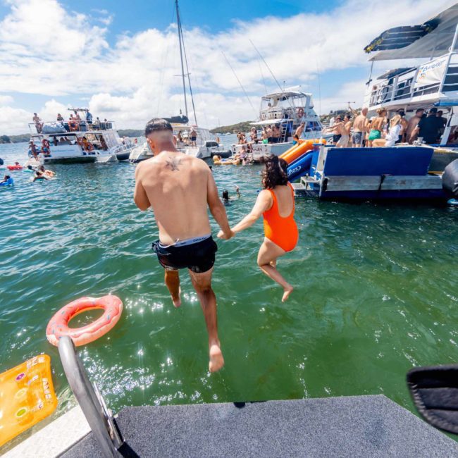 Two people are jumping off a boat into the water with multiple boats and people swimming and relaxing in the background under a clear blue sky, showcasing the lively atmosphere of a Sydney boat party hire event.