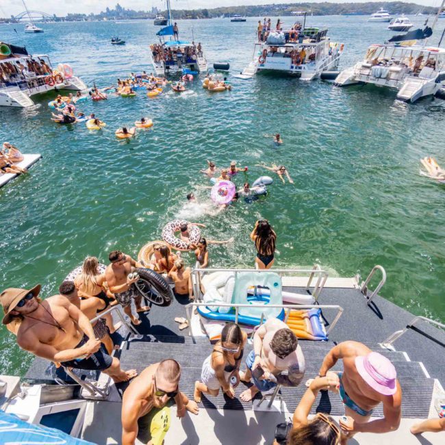 People enjoying a sunny day on a luxury yacht hire in Sydney, swimming, lounging on floats, and socializing. Several boats are anchored nearby with attendees on board and water toys in the water.