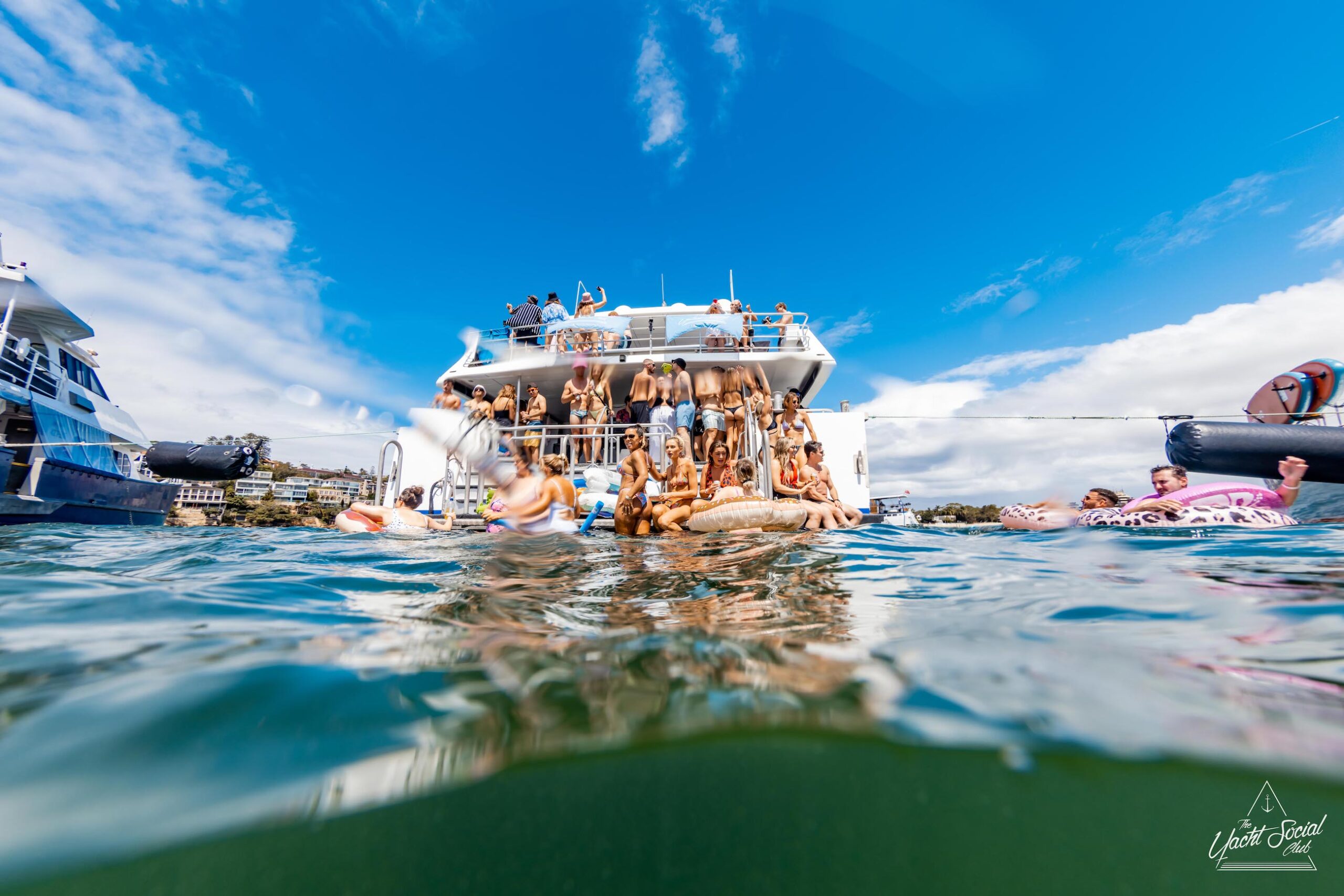 A group of people are on and around a luxury yacht on the water in Sydney Harbour. Some are sitting on the edge, while others are swimming or floating on inflatables. The sky is mostly clear with some clouds.
