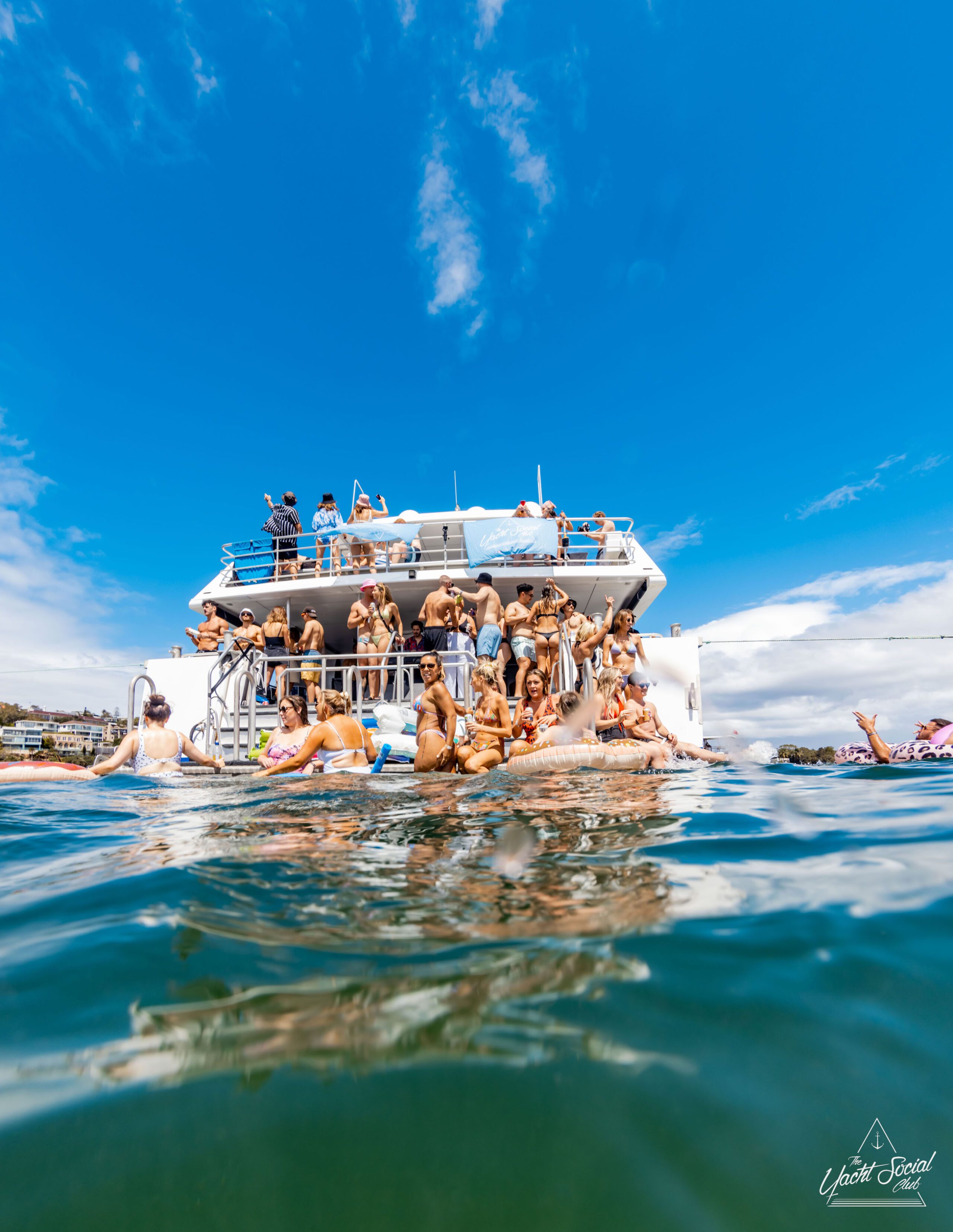 A group of people gather on a two-story luxury yacht, with some standing on the deck and others swimming in the water below, under a clear blue sky.