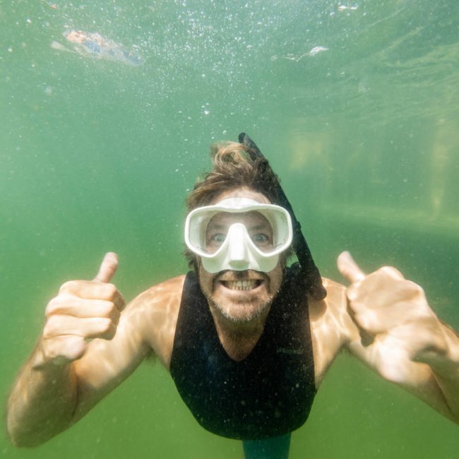 A person wearing a snorkel mask underwater gives a thumbs-up with both hands. The water, illuminated by the nearby Catamaran party Sydney, is greenish and slightly murky.