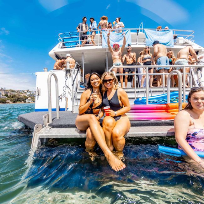 A group of people are on and around a catamaran party in Sydney. Some are sitting at the back of the boat, while others are swimming or relaxing in the water with pool noodles.