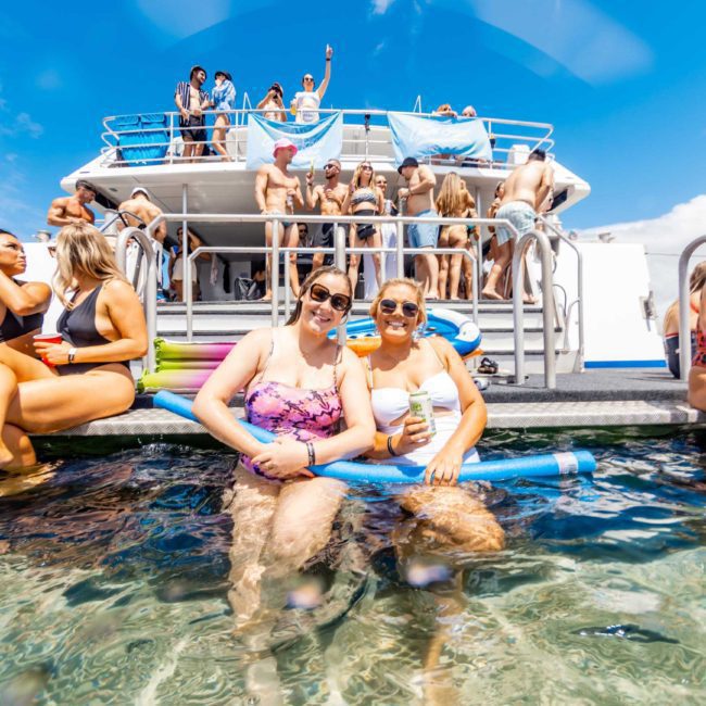 People enjoying a sunny day on a boat; some are sitting on the deck with their feet in the water, while others are gathered on the upper deck during a Catamaran party in Sydney.