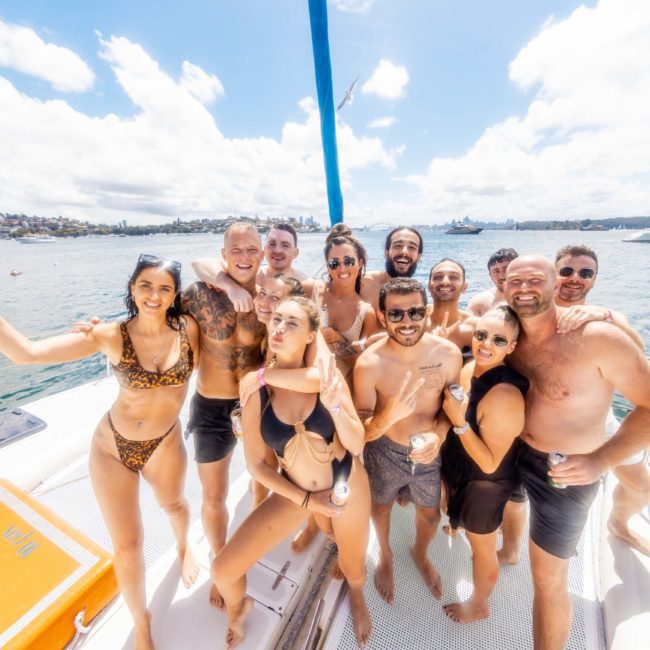 A group of people in swimwear smiling and posing for a photo on a boat under a sunny sky, with water and other boats in the background, enjoying their unforgettable DJ boat hire Sydney experience.
