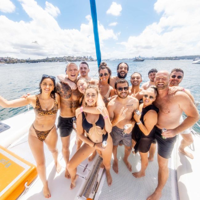 A group of people in swimwear smiling and posing on a boat under clear skies near the shoreline, with another boat in the background on the water during a private yacht charter Sydney Harbour.