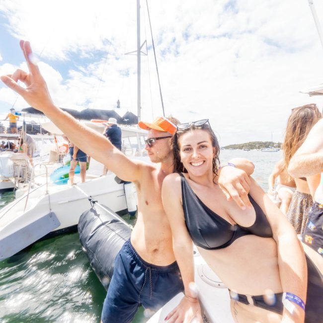 A group of people enjoying a sunny day on a luxury yacht hire Sydney, with one person pointing upwards and others smiling. The scene includes blue skies and other boats in the background.