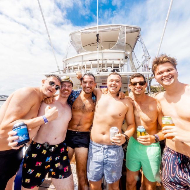 A group of six men in swimwear pose together on a boat, smiling and holding drinks. The background shows a clear sky and parts of other boats. Enjoy the ultimate experience with a private yacht charter Sydney Harbour, perfect for celebrations on the water.