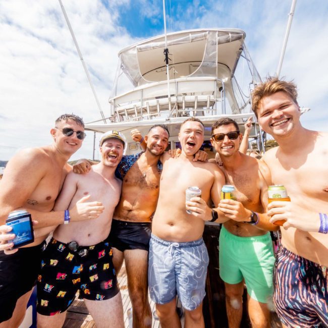 A group of seven men stand on a boat deck, smiling and holding drinks. They are enjoying a sunny day with a luxury yacht hire Sydney in the background.