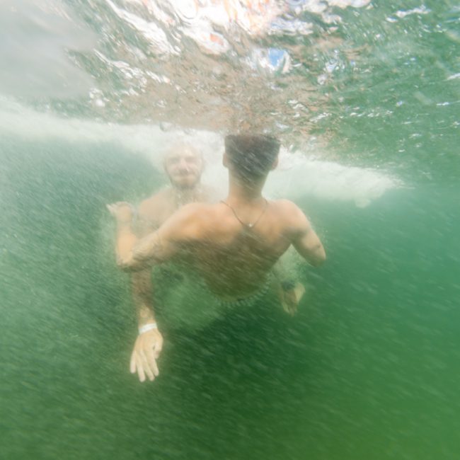 Two men are underwater in a swimming pool, facing each other, with one extending his arm towards the other. Picture yourself on a seamless Luxury yacht hire in Sydney, where every moment feels as weightless and serene as this.