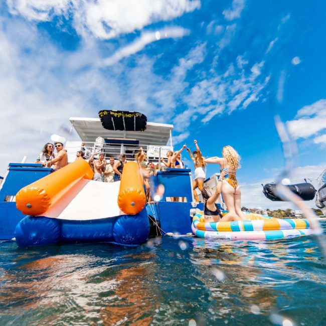 A group of people enjoying a sunny day on a luxury yacht with an inflatable slide and float. Some are on the upper deck, while others are on the float in the water. Another boat is visible in the background, perfect for a Corporate boat event or Sydney boat party hire.