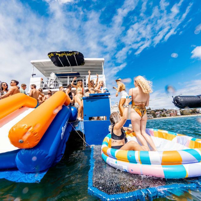 A group of people enjoy a sunny day on a luxury yacht hire in Sydney, complete with an inflatable slide and a colorful inflatable pool in the water.