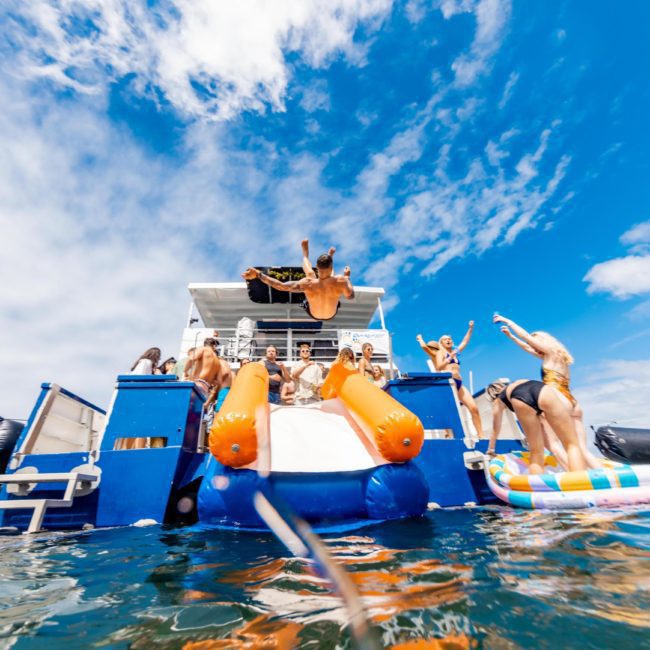 A group of people are gathered on a boat with an inflatable slide. One person is jumping into the water while others watch. There is another boat in the background, setting the scene for a lively catamaran party in Sydney Harbour.