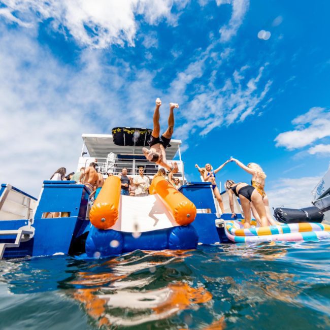 People enjoying a sunny day on a catamaran party in Sydney, complete with waterslides and a floating platform. A person is in mid-air, performing a flip off the boat while others watch and prepare to join in the water activities.