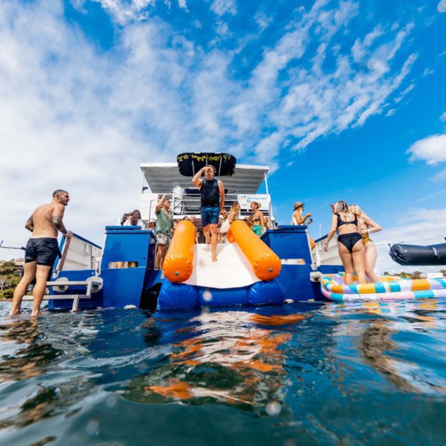 A person slides down an inflatable slide from a blue boat into the water, while others watch and a group stands on a nearby boat under a blue sky with scattered clouds. It's the perfect backdrop for those considering DJ boat hire Sydney or even planning a vibrant Catamaran party Sydney.