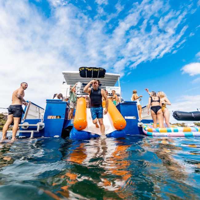 A person slides into the water from a houseboat while others stand or sit around, with several people on the deck of an adjacent boat. The sky is partly cloudy, perfect for a Sydney boat party hire on a private yacht charter in Sydney Harbour.