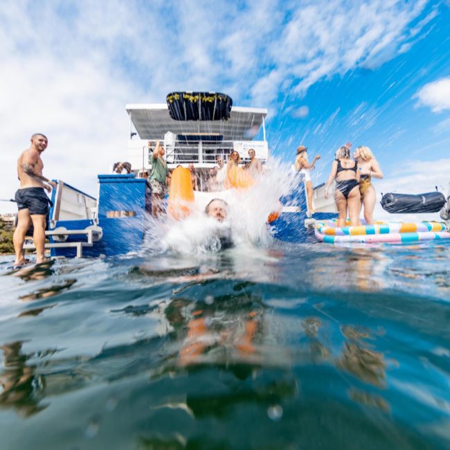 A person makes a big splash as they jump off the back of a boat into the water, with other people on the boat watching and some floating nearby on inflatables, enjoying a lively DJ boat hire Sydney experience.