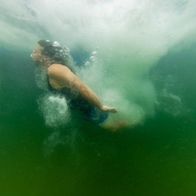A person swims underwater with bubbles trailing behind them in greenish water during a Sydney boat party hire.