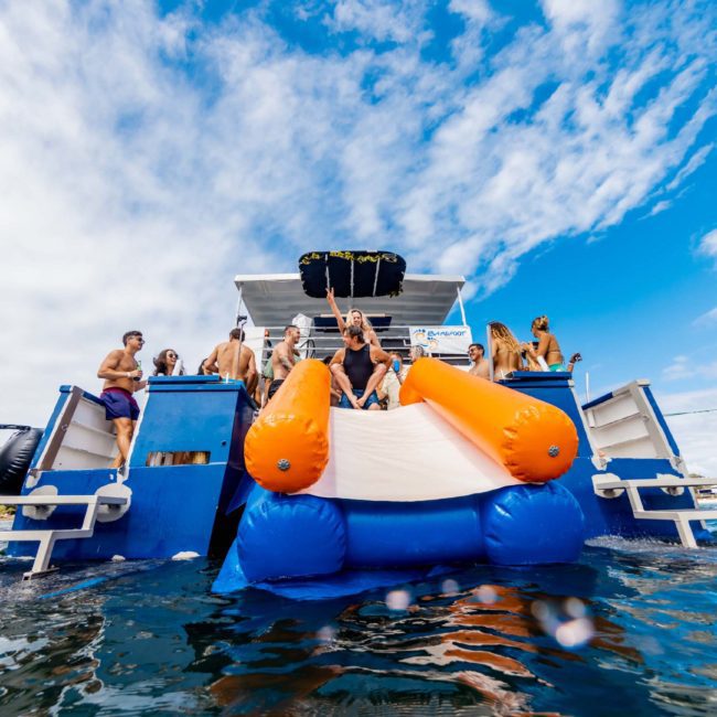 A group of people enjoy an inflatable water slide on a luxury yacht under a partly cloudy sky, surrounded by water with another boat in the background.