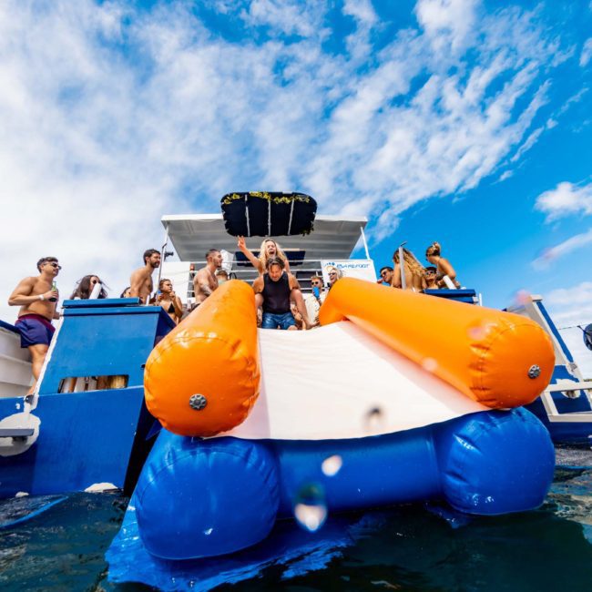 People are gathered on a boat, with one person sliding down an inflatable slide into the water. The sky is bright and partly cloudy—a perfect day for a catamaran party in Sydney.