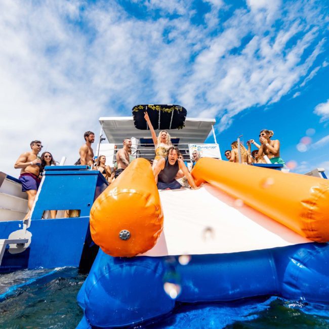 A group of people enjoy a Sydney boat party hire with a large inflatable slide into the water. One person is gracefully sliding while the others watch and snap photos. The sky is partly cloudy.