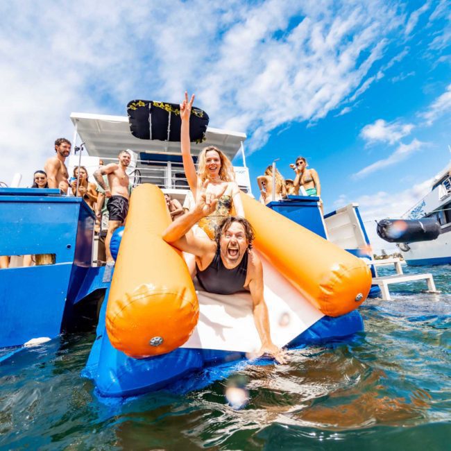 A person joyfully sliding down an inflatable slide from a catamaran party Sydney into the water, surrounded by a cheerful crowd under a clear blue sky.
