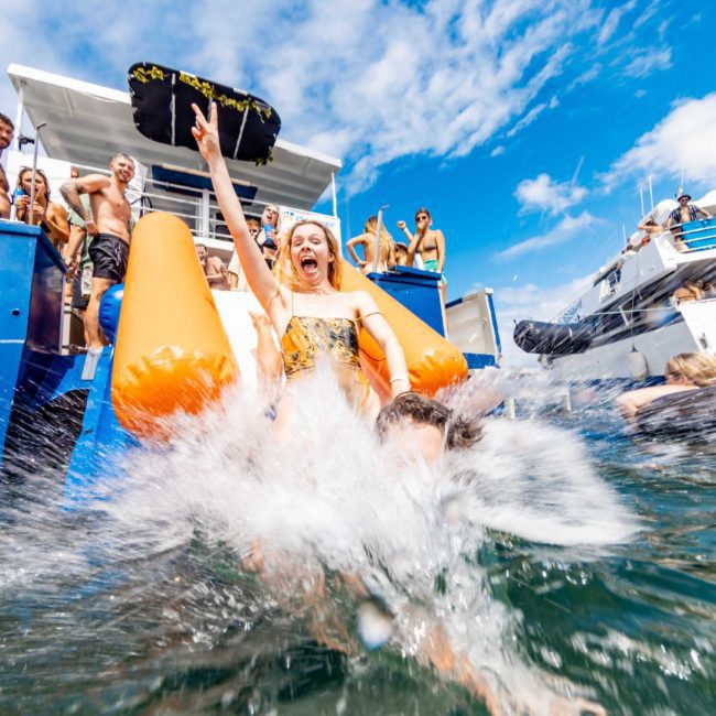 A woman slides down an orange inflatable slide into the water from a boat as others watch, with additional boats in the background under a blue sky, capturing the vibrant scene of a private yacht charter Sydney Harbour.