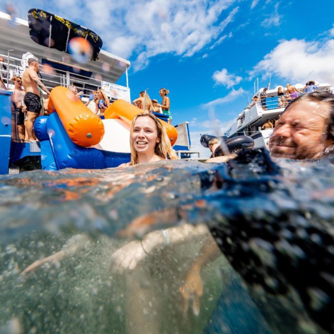 People enjoy swimming near boats in bright, sunny weather, with inflatables and various activities on board. For an unforgettable experience, consider a Sydney boat party hire or a catamaran party in Sydney.